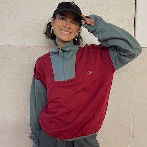 Könnte beinhalten: Ein weinrot-teal farbblockierter Sweatshirt mit einem Viertel-Reißverschlusskragen. Das Sweatshirt hat tealfarbene Ärmel und einen tealfarbenen Kragen. Ein kleines Logo ist auf der Vorderseite sichtbar. Die Person trägt eine schwarze Baseballkappe.