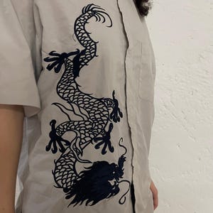 Könnte beinhalten: Hellgraues Hemd mit kurzen Ärmeln und einem dunkelblauen Drachenmotiv, das auf die Vorderseite gestickt ist. Das Hemd hat eine Knopfleiste und eine Tasche auf der linken Seite.