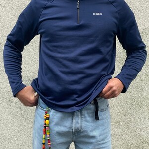 Op de afbeelding: Marineblauw shirt met lange mouwen, een kraag met ritssluiting en de merknaam "odlo" in het wit. Het shirt wordt gecombineerd met een lichtblauwe jeans en een kleurrijk kralenaccessoire.