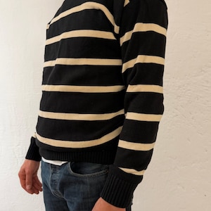 Könnte beinhalten: Ein schwarz-cremefarbener gestreifter Pullover. Der Pullover hat einen Rundhalsausschnitt und lange Ärmel mit gerippten Bündchen. Die Streifen sind horizontal und gleichmäßig angeordnet. Die Person trägt eine blaue Jeans.