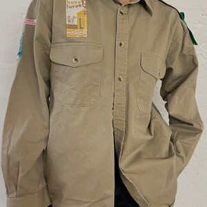 Könnte beinhalten: Khakifarbene Button-Down-Hemd mit zwei Brusttaschen und einem Kragen. Das Hemd hat Aufnäher mit Text und Illustrationen. Grünes Band an der Schulter befestigt. Ein Vintage-Casual-Stil.