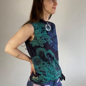 Könnte beinhalten: Ärmelloses schwarzes Oberteil mit blau-grünem floralem Spitzendesign. Das Oberteil hat einen hohen Kragen und einen silbernen Anhänger. Das Model trägt blaue Jeans mit rosa und lila Akzenten auf den Gesäßtaschen.