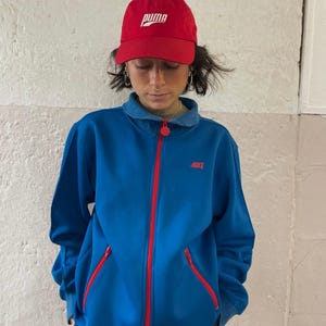 Könnte beinhalten: Eine rote Puma-Baseballkappe mit weißem Schriftzug wird mit einer blauen Nike-Jacke mit Reißverschluss getragen. Die Jacke hat einen roten Reißverschluss und rote Akzente an den Taschen. Das Nike-Logo ist rot gestickt. Das Outfit ist lässig und sportlich.