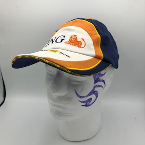Könnte beinhalten: Eine marineblaue, orangefarbene und weiße Baseballkappe mit dem Wort "ING" und einem Löwenlogo. Der Schirm hat das Wort "RENAULT" wiederholt aufgedruckt. Die Kappe befindet sich auf einem weißen Schaufensterkopf.