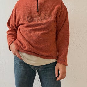 Könnte beinhalten: Rostfarbener Fleece-Pullover mit Stehkragen und schwarzem Reißverschluss. Das Wort "GAGE" ist auf die Brust gestickt. Der Pullover wird mit einer blauen Jeans kombiniert. Der Stil ist lässig und bequem.