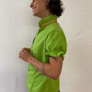 Può includere: Camicetta verde brillante con collo alto a balze e maniche corte a sbuffo. Il colletto è rifinito con un motivo a zig-zag rosa. La camicetta ha una chiusura a bottoni ed è abbinata a una gonna fantasia bianca e nera.