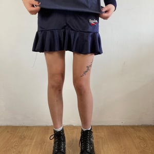 Könnte beinhalten: Marineblauer Sportrock mit Rüschensaum und Reebok-Logo. Der Rock wird mit schwarzen Schnürstiefeln getragen. Die Person trägt ein marineblaues Oberteil.