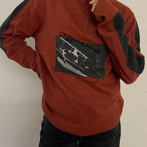 Könnte beinhalten: Rostfarbener Pullover mit Stehkragen und Viertelreißverschluss. Der Pullover hat dunkelgraue Akzente an den Ärmeln und einen rechteckigen Aufnäher mit dem Text "NORTHLAND PRO" und einer Grafik.