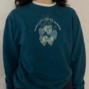 Op de afbeelding: Teal sweatshirt met witte bies en geborduurde harten. De tekst "Grandma's Love is tied with Heartstrings" is rond de harten gebogen. De harten hebben bloemen- en patroonontwerpen.