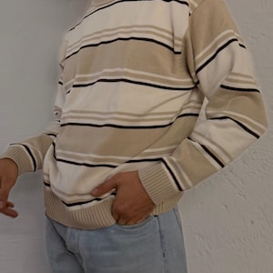 Könnte beinhalten: Ein beige-weiß gestreifter Pullover mit schwarzen Akzentstreifen. Der Pullover hat einen gerippten Kragen, Bündchen und Saum. Die Person trägt eine hellblaue Jeans. Der Pullover ist ein lässiges Kleidungsstück für jeden Tag.