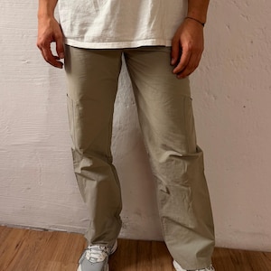 Könnte beinhalten: Khakifarbene Cargohose mit Seitentaschen und lockerer Passform. Die Hose wird mit einem weißen T-Shirt und weißen und grauen Sportschuhen getragen. Das Bild ist vor einer weißen Wand aufgenommen.