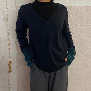 Könnte beinhalten: Eine marineblaue Strickjacke mit V-Ausschnitt und Knopfverschluss. Die Ärmel sind lang und die Manschetten grün und blau gestreift. Die Strickjacke wird über einem schwarzen Rollkragenpullover getragen. Die Person trägt auch eine graue Hose.