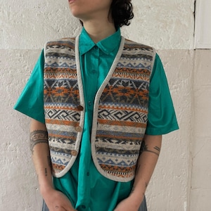 Op de afbeelding: Een vest met patroon met een crèmekleurige bies en houten knopen. Het vest heeft een geometrisch ontwerp in tinten oranje, blauw en grijs. Het wordt gedragen over een turquoise button-down overhemd en een blauwe broek.
