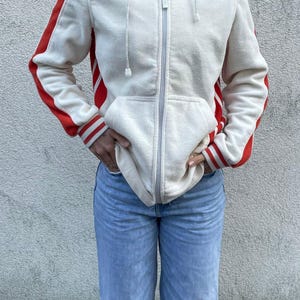 Könnte beinhalten: Cremefarbener Kapuzenpullover mit roten Streifen an den Ärmeln und Bündchen. Der Hoodie hat eine Kapuze mit Kordelzug und eine Vordertasche. Die Person trägt eine hellblaue Jeans.