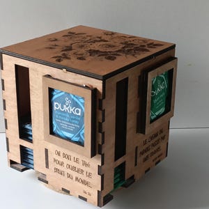 Peut inclure: Boîte à thé en bois avec un motif floral découpé au laser sur le couvercle. La boîte comporte des compartiments pour les sachets de thé, avec des fenêtres encadrées affichant les étiquettes de la marque de thé. Du texte en français est visible.
