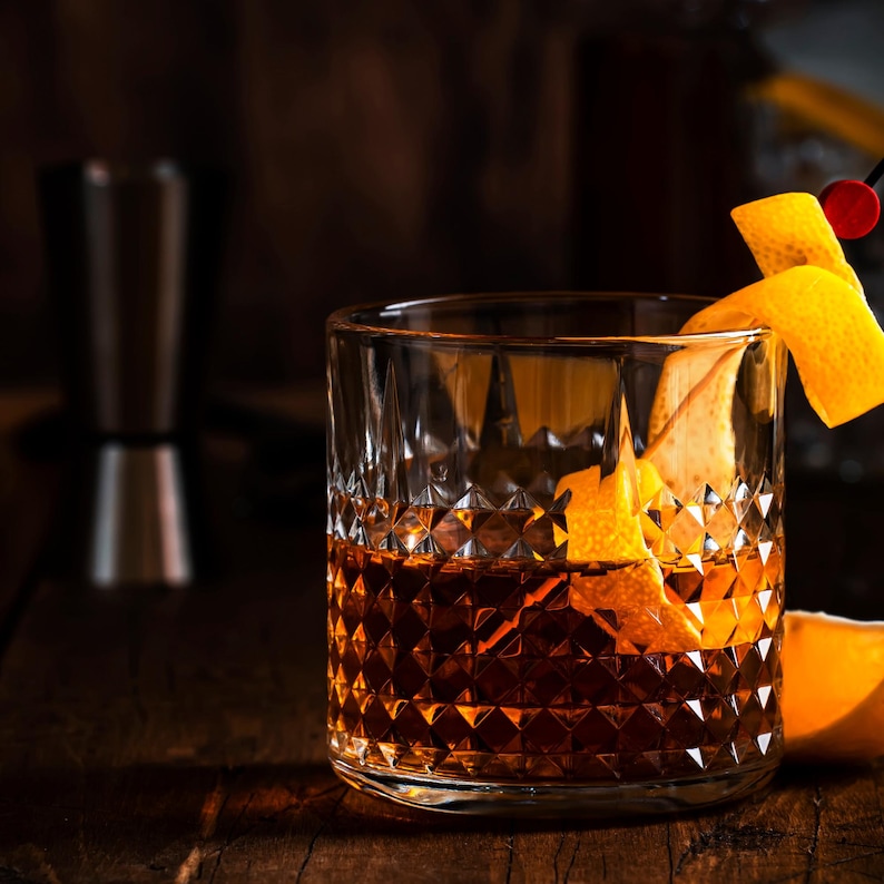 May include: A rocks glass filled with a dark amber cocktail, garnished with an orange peel and a cherry. The glass has a diamond pattern. A stainless steel jigger is in the background, along with a wooden surface.