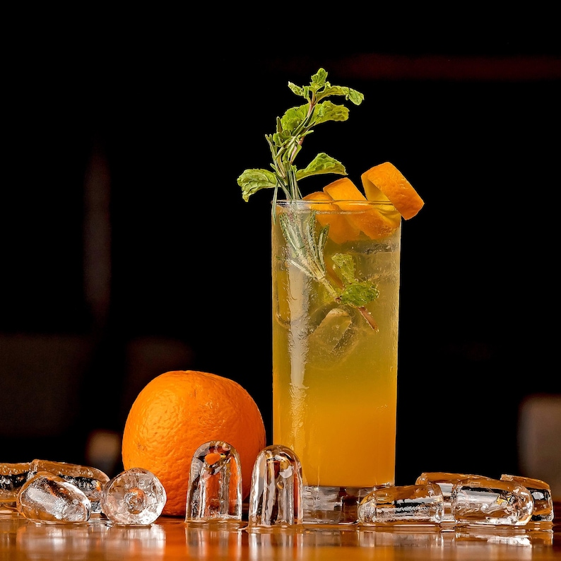May include: A tall glass of orange-colored cocktail garnished with orange slices and sprigs of mint. An orange sits next to the glass, with ice cubes scattered around. The drink is set against a dark background.