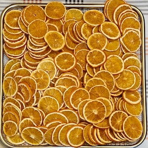 May include: A metal tray filled with numerous dried orange slices. The slices are a warm, golden-orange color with a lighter-colored rind. The arrangement is dense, with slices overlapping, creating a textured surface. The tray sits on a checkered surface.