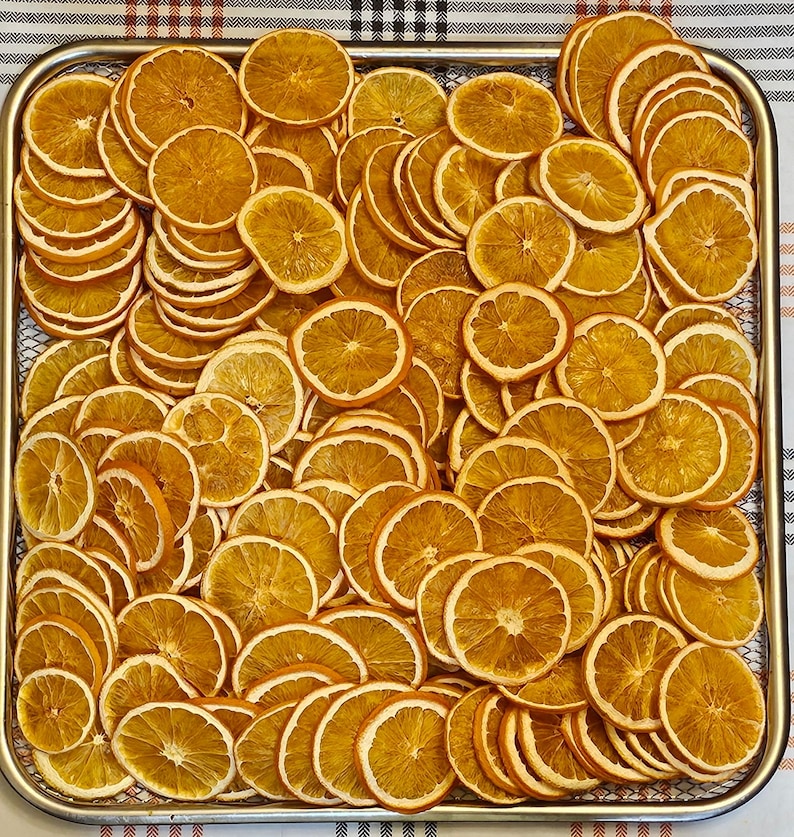 May include: A metal tray filled with numerous dried orange slices. The slices are a warm, golden-orange color with a lighter-colored rind. The arrangement is dense, with slices overlapping, creating a textured surface. The tray sits on a checkered surface.