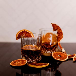 May include: Two crystal glasses filled with a dark liquid, garnished with dried orange slices. The glasses sit on a reflective surface, with additional orange slices scattered around. The background is a neutral, textured wall.