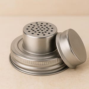 Op de afbeelding: Een zilverkleurige deksel voor een weckpot met een geperforeerde bovenkant en een apart, kleiner deksel. De deksel is ontworpen om op een standaard weckpot te passen, met een schroefdraad voor een veilige bevestiging. De bovenkant heeft meerdere kleine gaatjes.