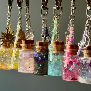 Könnte beinhalten: Eine Sammlung kleiner Glasflaschen mit Korken, gefüllt mit Glitzer und Anhängern. Jede Flasche hängt an einer silbernen Kette und einem Verschluss. Zu den Anhängern gehören eine Sonne, ein Smiley und ein Mond. Die Flaschen enthalten verschiedene Farben von Glitzer.