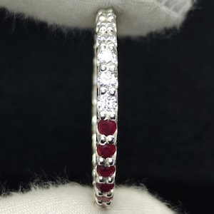 May include: A thin silver eternity band featuring a row of alternating round-cut clear white stones and deep red rubies set in a metallic frame. The ring is held vertically, showcasing the continuous pattern of gemstones along the entire circumference.