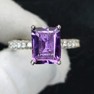 May include: A silver ring with a large, rectangular, purple gemstone. The gemstone is the focal point, held by four prongs. The band is embellished with smaller, clear gemstones. The ring is shown against a white and black backdrop.