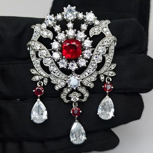 Op de afbeelding: Een sierlijke zilveren broche met een grote, vierkante, rode edelsteen in het midden, omringd door kleinere, heldere stenen. De broche heeft een bloemmotief met hangende peervormige en ronde edelstenen. Een luxueus sieraad.