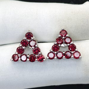 May include: A pair of triangular stud earrings. Each earring is composed of multiple round, deep red gemstones set in a silver metal. The earrings are displayed on a white fabric surface.