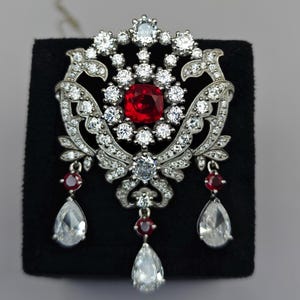 May include: An elaborate silver brooch, featuring a central square red gemstone encircled by many small, clear gemstones. The brooch has a floral design with hanging teardrop-shaped clear gemstones and smaller red gemstones.