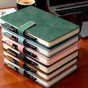 May include: A stack of notebooks in various colors, including green, blue, pink, black, brown, and gray. Each notebook has a pen secured by a matching colored strap and a gold-colored clasp. The notebooks are stacked on a wooden surface.