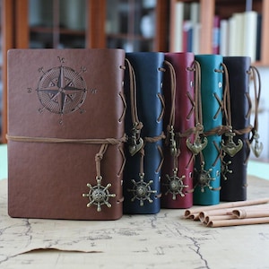 Op de afbeelding: Een verzameling lederen journals in verschillende kleuren: bruin, marineblauw, bordeauxrood, blauwgroen en zwart. Elk journal heeft een kompasroosontwerp en een decoratieve bedel. De journals zijn vastgebonden met een leren koord.