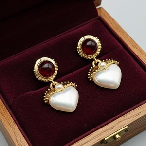 Peut inclure: Boucles d'oreilles dorées avec des pendentifs en forme de cœur en perle. Chaque boucle d'oreille comporte une pierre rouge et une petite perle. Les boucles d'oreilles sont présentées dans un coffret en bois avec un intérieur en velours bordeaux.