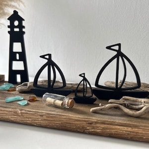 Könnte beinhalten: Schwarze Holzfiguren von Segelbooten und einem Leuchtturm auf einem Treibholzsockel. Dekorative Elemente sind Meerglas, eine kleine Glasflasche mit einer Schriftrolle und getrocknete Äste. Der Leuchtturm ist ca. 15 cm hoch.