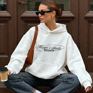 Puede incluir: Sudadera con capucha blanca con el texto "Meggie's Studio Mockup" en fuente negra. La sudadera tiene un bolsillo delantero y se combina con vaqueros oscuros y zapatillas blancas. Un bolso de hombro marr&oacute;n y gafas de sol completan el look.