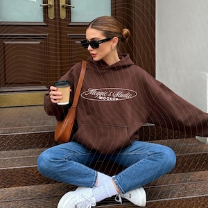 May include: A brown hoodie with the text "Meggies Studio Mockup" in a white script font. The hoodie has a front pocket and is worn with blue jeans and white sneakers. The person is wearing black sunglasses and holding a coffee cup.