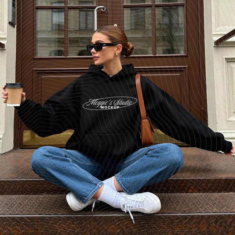 Puede incluir: Una sudadera con capucha negra con el texto "Meggie's Studio Mockup" en blanco, combinada con vaqueros azules y zapatillas blancas. La persona sostiene una taza de caf&eacute; y lleva gafas de sol. Tambi&eacute;n se ve un bolso de hombro marr&oacute;n.