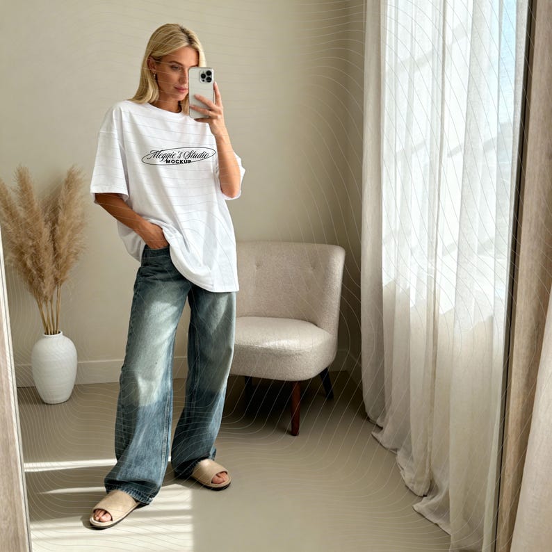 May include: White oversized t-shirt with the text "Maggie's Studio Mockup" in black script. The model is wearing blue jeans and beige slides. The background includes a white vase with dried plants, a beige chair, and a white curtain.