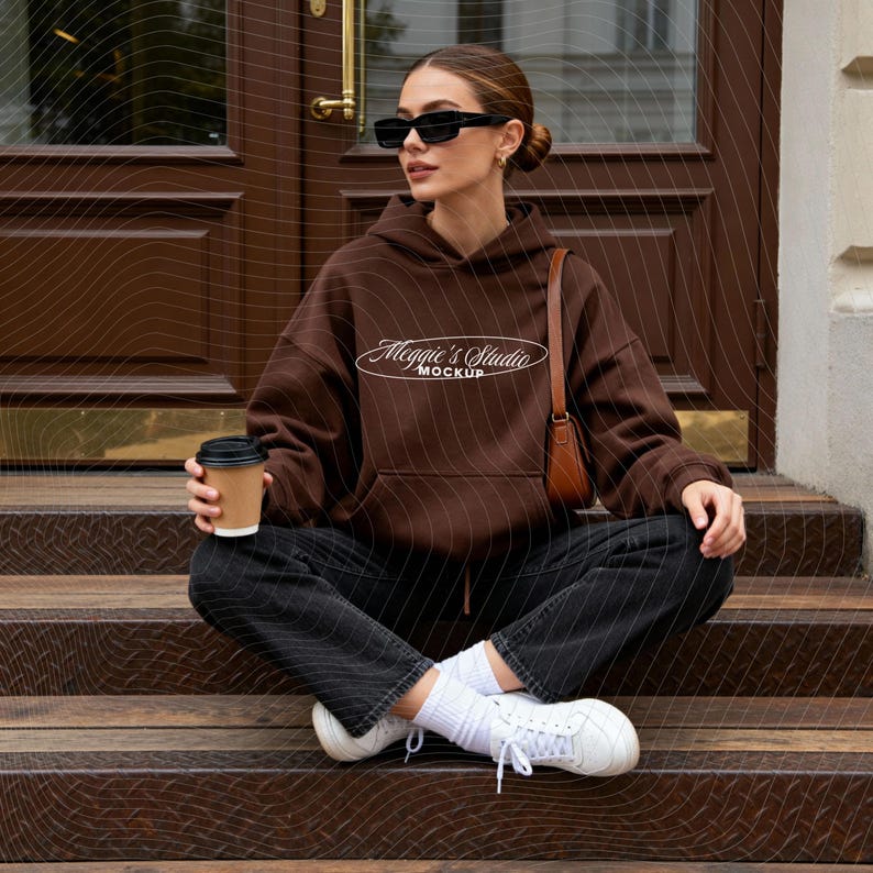 May include: A brown hoodie with the text "Maggie's Studio Mockup" is worn by a person sitting on wooden steps. The person is wearing black jeans, white sneakers, and sunglasses, and holding a coffee cup. A brown purse is over the shoulder.