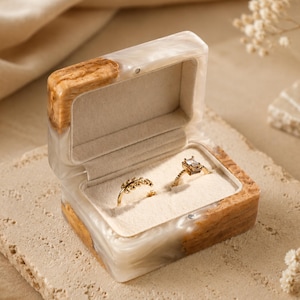 Könnte beinhalten: Geöffnete Holz-Ringbox mit zwei goldenen Ringen im Inneren. Die Box hat eine hellfarbene, marmorierte Harzaußenseite mit einem Holzrahmen. Die Ringe liegen auf einem cremefarbenen Samtinnenraum. Die Ringe haben einen Diamanten und ein Blumenmuster.