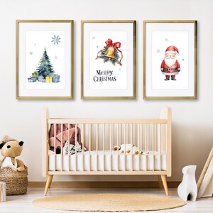 Könnte beinhalten: Drei gerahmte Weihnachtsdrucke in Aquarell. Die Drucke zeigen einen Weihnachtsbaum, eine Glocke mit roter Schleife und den Weihnachtsmann. Die Drucke sind in goldenen Rahmen und werden über einem Kinderbett in einem Kinderzimmer ausgestellt.