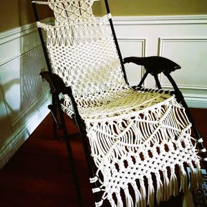 Può includere: Sedia pieghevole in stile macramè. La seduta e lo schienale sono in tessuto color crema con frange. Il telaio nero sostiene la sedia, progettata per il relax all'aperto. Comoda.