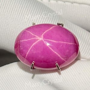 May include: A vibrant, oval-shaped, pink gemstone with a star-like pattern. The gemstone is set in a silver metal mounting. The gem is displayed on a white fabric background.