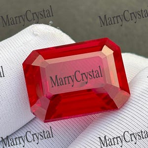 Könnte beinhalten: Ein leuchtend roter Edelstein im Smaragdschliff mit facettierter Oberfläche. Der Edelstein liegt auf einem weißen, strukturierten Stoff. Der Text "MarryCrystal" ist auf dem Edelstein zu sehen und wiederholt sich im Hintergrund in schwarzer Schrift.