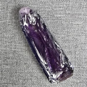 Puede incluir: Una punta de cristal de amatista pulida y alargada con un tono púrpura intenso. La superficie de la piedra es lisa y reflectante, mostrando variaciones naturales en la intensidad del color y la claridad interna a lo largo de su forma.
