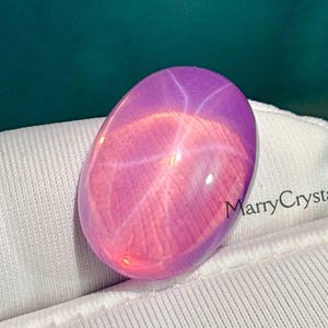 Puede incluir: Una gema ovalada pulida con efecto estrella. La gema presenta un degradado de tonos rosa y morado, con un prominente patrón de estrella que irradia desde el centro. La gema se sostiene en una tela blanca.