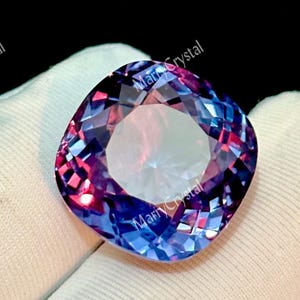 Pode incluir: Uma pedra preciosa de corte almofada exibindo um gradiente de cores mutáveis em tons de roxo profundo, azul vibrante e magenta. A pedra facetada apresenta uma superfície brilhante e reflexiva com bordas nítidas e um centro transparente.