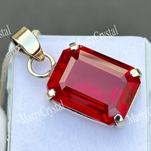May include: A rectangular, faceted red gemstone pendant set in a silver-toned metal prong setting. The pendant is attached to a silver-toned chain and rests on a white square display base. The text MarryCrystal is visible in the background.
