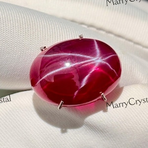 May include: A large, oval-shaped, deep red gemstone with a six-ray star effect. The gem is set in a silver-colored metal. The image is of a close-up of the gemstone, showcasing its polished surface and star pattern.